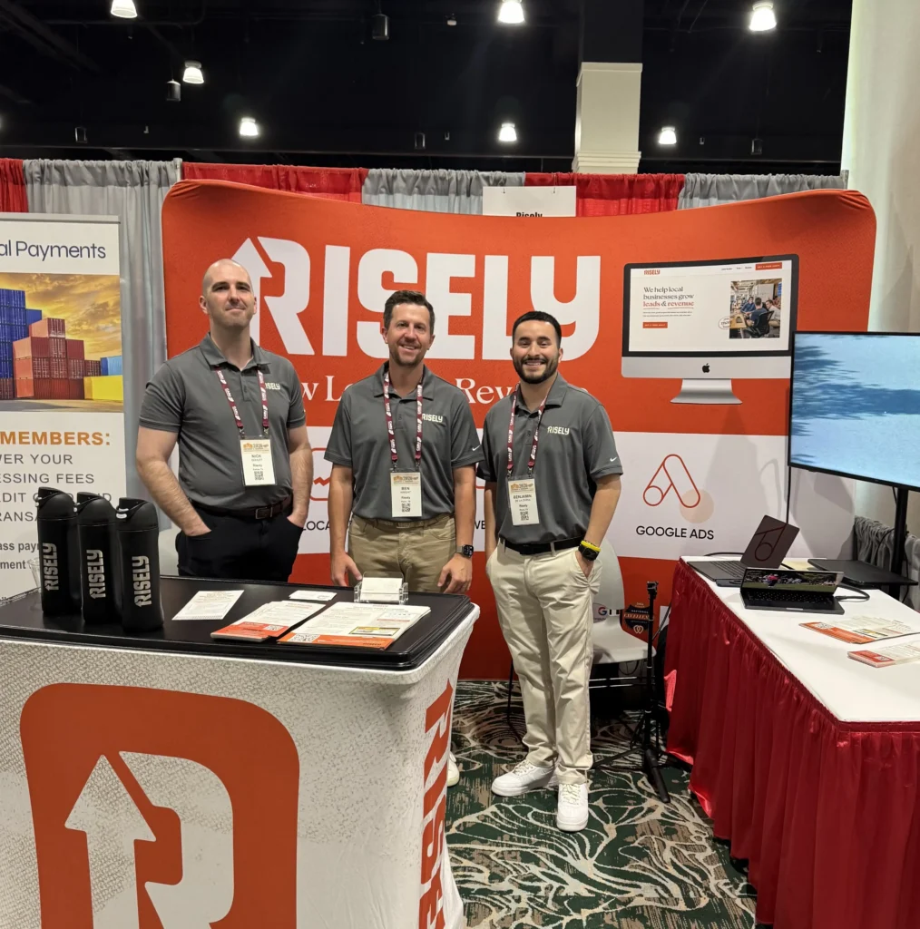 Three Risely team members standing at their orange-branded trade show booth featuring Local SEO, Web Design, and Google Ads services at a 2026 industry conference.