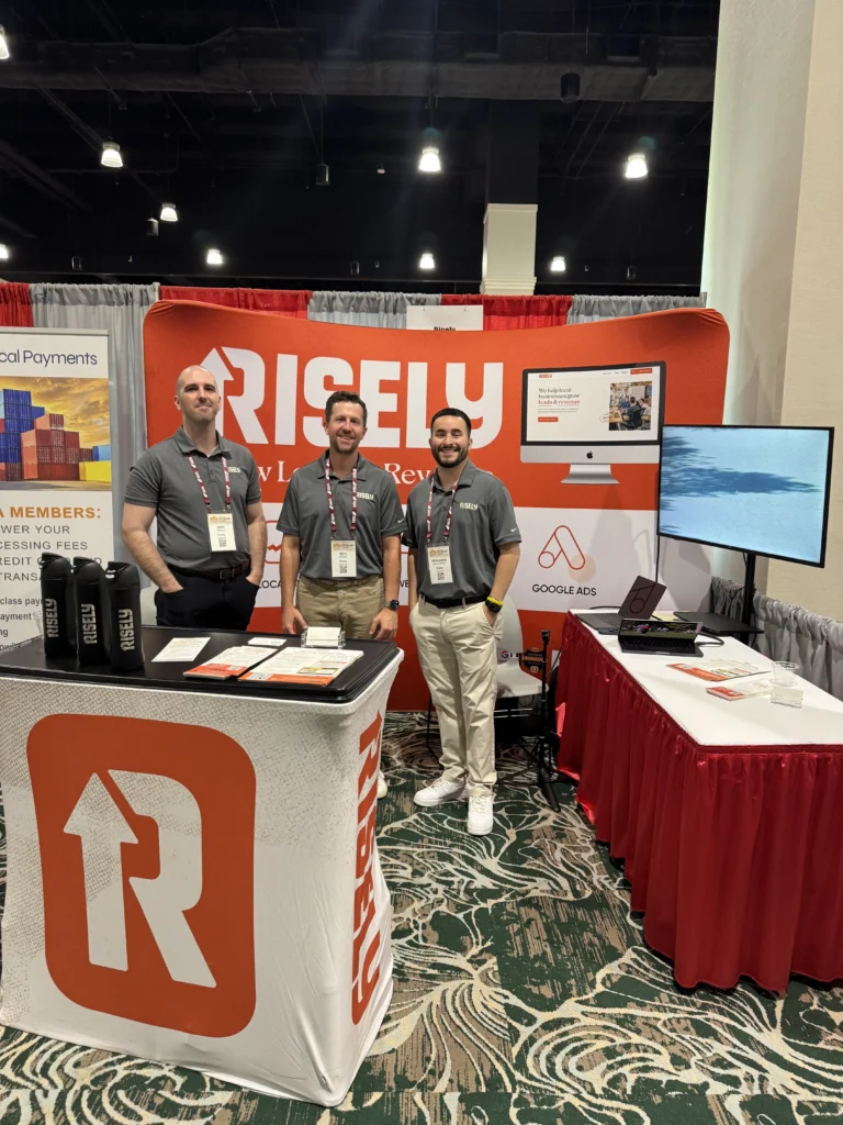 Three Risely team members standing at their orange-branded trade show booth featuring Local SEO, Web Design, and Google Ads services at a 2026 industry conference.