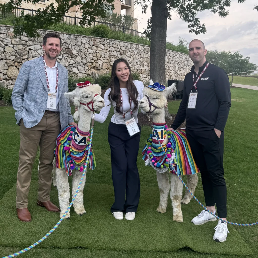Three Risely team members posing outdoors with two decorated alpacas wearing colorful blankets and small sombreros at a conference networking event.